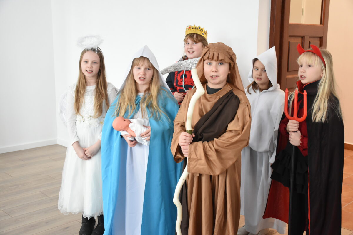 Sześcioro dzieci stoi przed obiektywem, przebranych w kostiumy jasełkowe: diabeł z trójzębem, śmierć w białej szacie, król Herod w koronie, anioł z aureolą, Maryja z lalką–Dzieciątkiem oraz pasterz z laską.