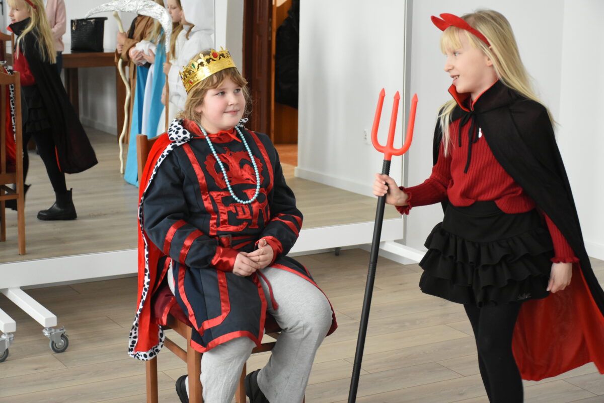 Dwoje dzieci w kostiumach jasełkowych stoi w sali: jedno siedzi na krześle przebrane za króla Heroda w złotej koronie i czerwono-czarnym stroju, a drugie jako diabeł z czerwonymi rogami trzyma trójząb i zwraca się do niego