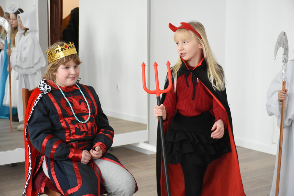 Dwoje dzieci w kostiumach jasełkowych stoi w sali: jedno siedzi na krześle przebrane za króla Heroda w złotej koronie i czerwono-czarnym stroju, a drugie jako diabeł z czerwonymi rogami trzyma trójząb i ostrzega króla.