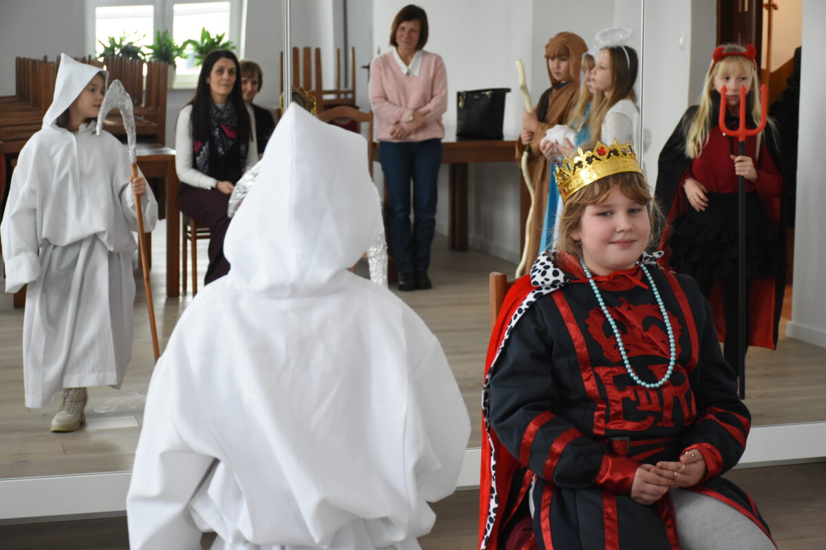 Dwoje dzieci w kostiumach jasełkowych stoi w sali: jedno siedzi na krześle przebrane za króla w złotej koronie i czerwono-czarnym stroju, a drugie jako śmierć w białej szacie z kosą i zwraca się do niego.