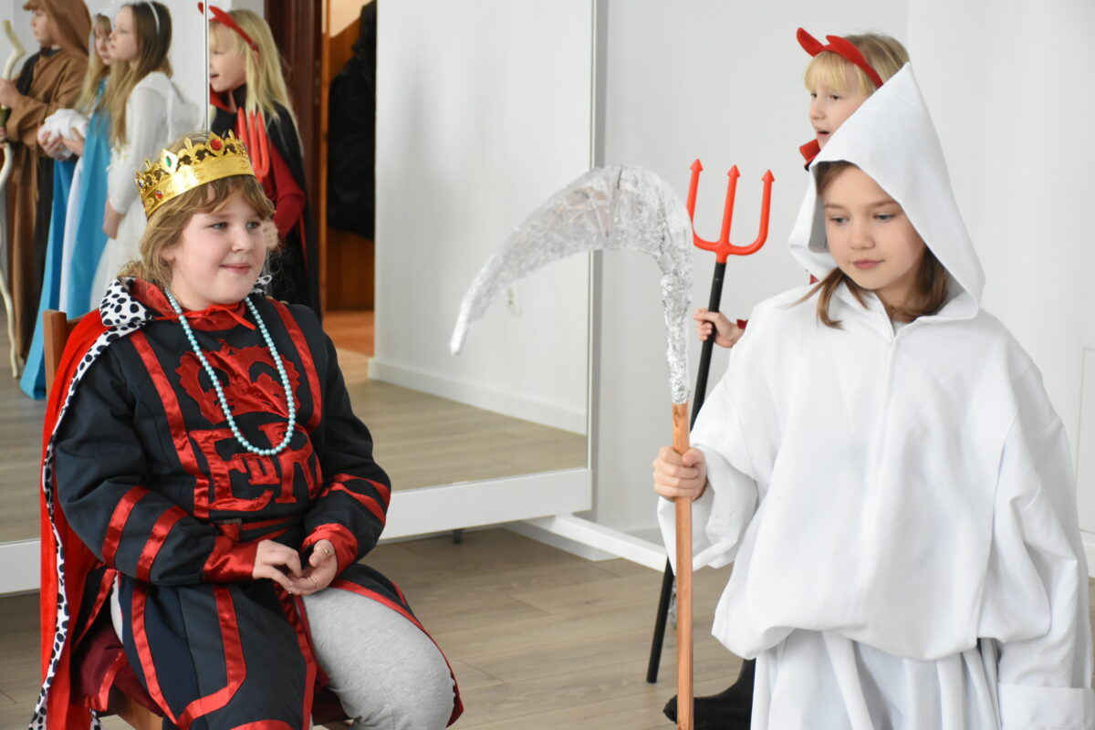 Dwoje dzieci w kostiumach jasełkowych stoi w sali: jedno siedzi na krześle przebrane za króla w złotej koronie i czerwono-czarnym stroju, a drugie jako śmierć w białej szacie z kosą i grozi że odbierze życie Herodowi