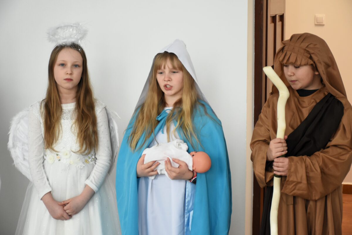 Trzech dzieci w kostiumach jasełkowych stoi przed objektywem: anioł z aureolą, Maryja z lalką–Dzieciątkiem oraz pasterz z laską składali życzenia