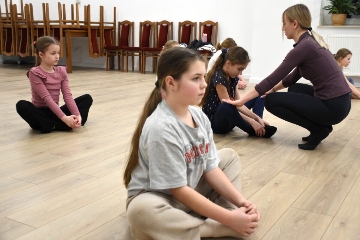Dziewczyny siedzą na podłodze na pośladkach, z nogami zgiętymi przed sobą, ale nie po turecku. Stopy są złączone ze sobą – stopa do stopy – a kolana rozchodzą się na boki