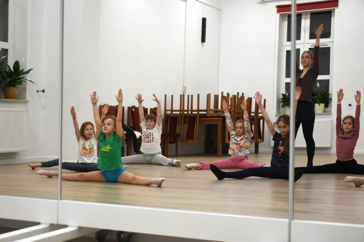 Dziewczyny siedzą rozłóżone szeroko wyprostowane nogi i rozciągają się do góry