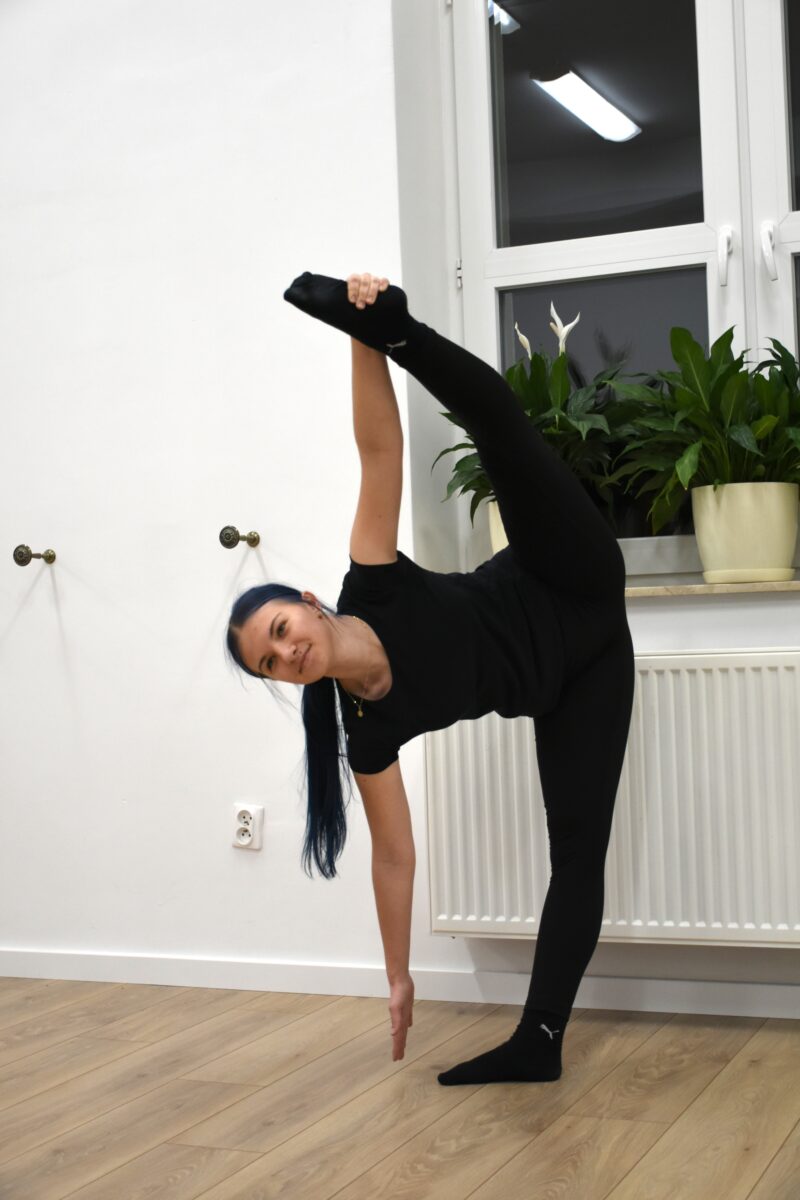 Dziewczyna jedna z uśmiechem pokazuje jak ładnie szpagat robi na stojąco na jedną nogę