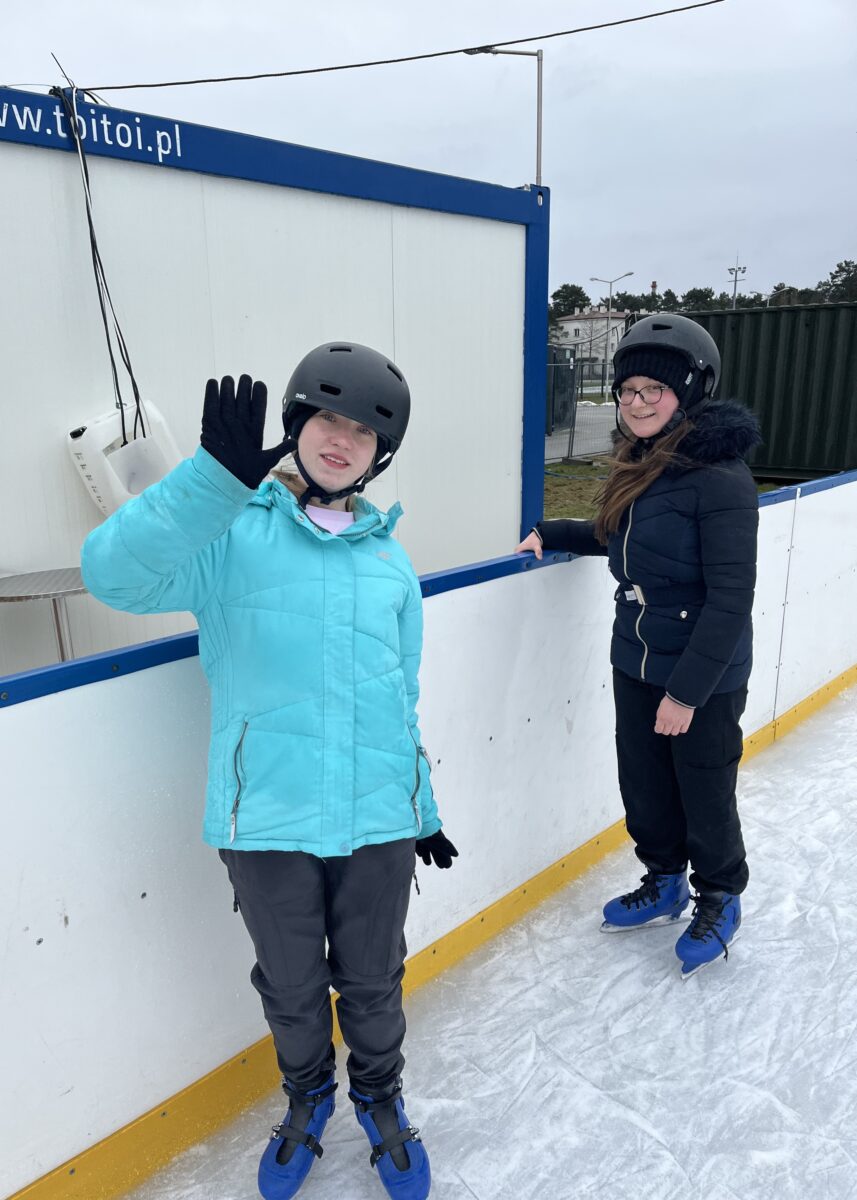 Sara macha ręką z uśmiechem do kamery i Sandra trzyma się barierki